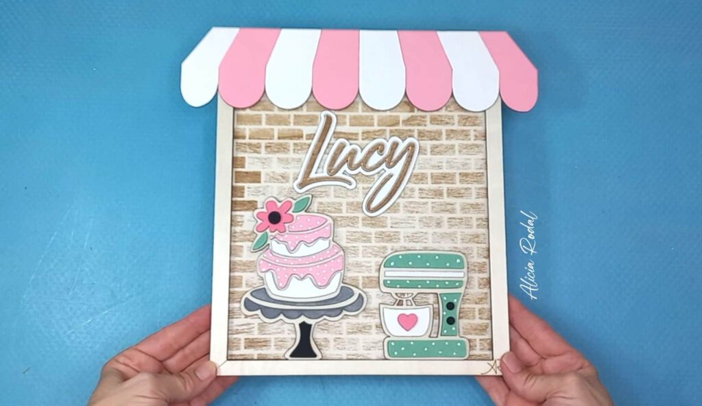 Hoy quiero compartir contigo el proceso detrás de una de mis creaciones más especiales: un cuadro de madera diseñado, cortado, grabado y pintado a mano, que representa una pequeña tienda u obrador de repostería. Esta pieza, pensada como regalo personalizado, forma parte del catálogo de mi tienda de arte y regalos personalizados, Alicia Rodal.